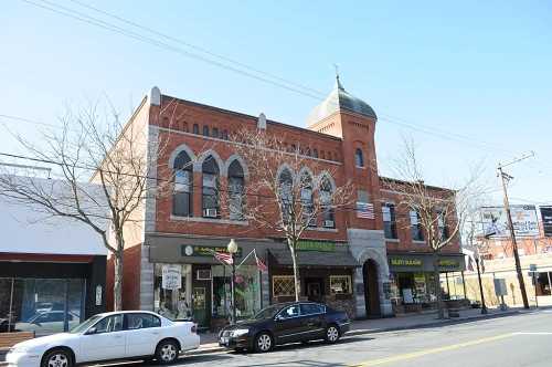 Historic brick building with arched windows, a dome, and storefronts, flanked by trees and parked cars on a street.