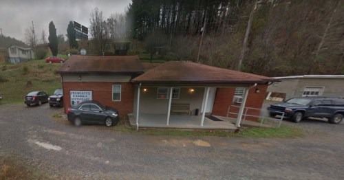 A small brick building with a porch, surrounded by cars, set in a rural area with trees in the background.