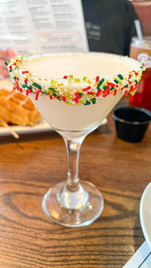 A festive cocktail in a glass with a colorful sprinkles rim, set on a wooden table with food in the background.
