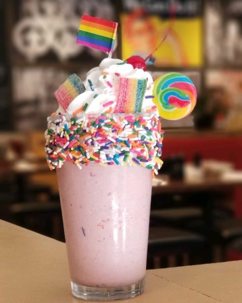 A colorful milkshake topped with whipped cream, rainbow sprinkles, a cherry, and festive decorations, including a rainbow flag.