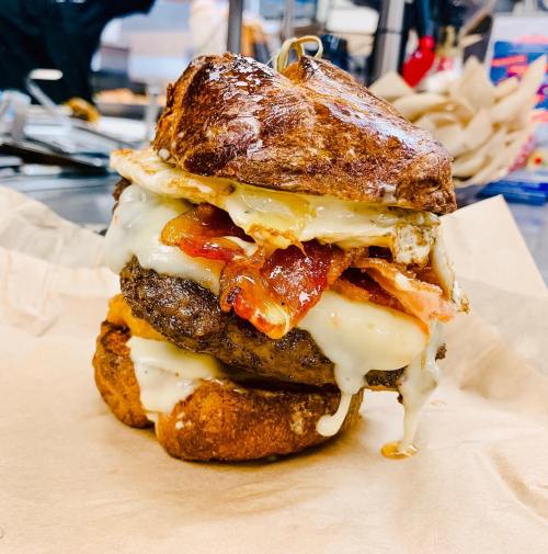 A towering burger with a beef patty, melted cheese, bacon, and a fried egg, served on a toasted bun.