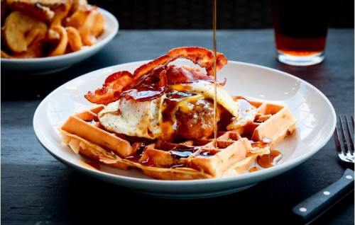 A plate of waffles topped with a fried egg, bacon, and syrup, with a drink in the background.