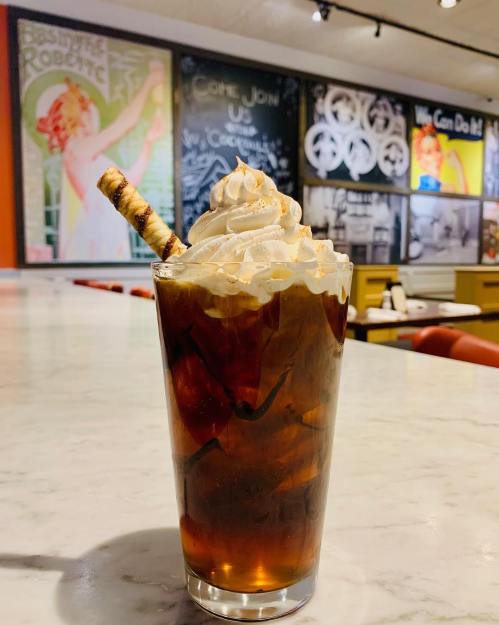 A glass of iced coffee topped with whipped cream and a decorative straw, set against a colorful mural backdrop.