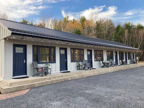 A row of modern motel rooms with blue doors, outdoor seating, and string lights, set against a wooded backdrop.
