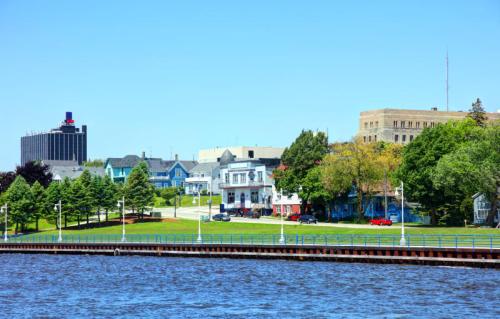 A scenic view of a riverfront with colorful houses, green trees, and buildings in the background under a clear blue sky.