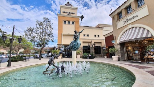 A fountain with a statue of a woman and children, surrounded by shops and trees in an outdoor shopping area.