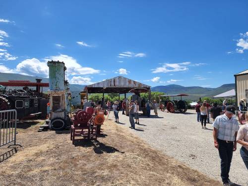 A sunny outdoor scene at a vintage machinery exhibit, with people walking among old tractors and equipment under a shelter.