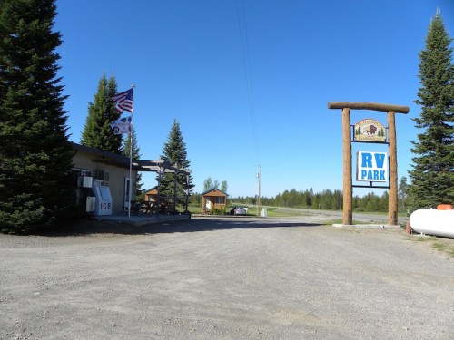 Entrance to an RV park with a large sign, American flag, and trees under a clear blue sky. Gravel road visible.
