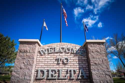 A stone welcome sign for Delta, featuring flags and a clear blue sky in the background.