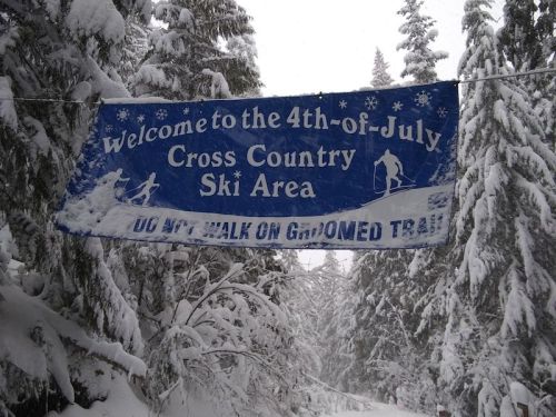 A snowy landscape with a banner reading "Welcome to the 4th-of-July Cross Country Ski Area" hanging between trees.