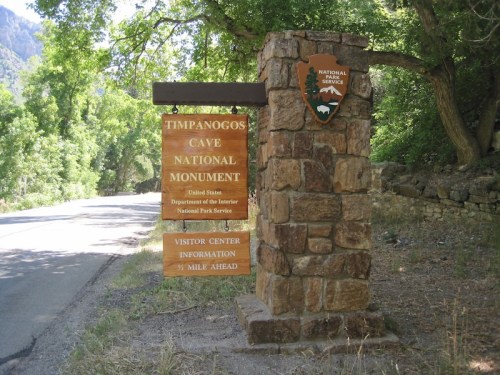 Sign for Timpanogos Cave National Monument, featuring visitor center information, surrounded by greenery.