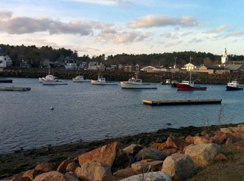 A serene harbor scene with several boats anchored, rocky shoreline, and a small town in the background under a cloudy sky.