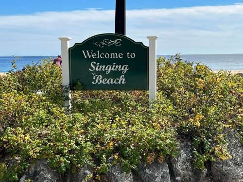 A sign reading "Welcome to Singing Beach" surrounded by greenery, with a beach and ocean in the background.