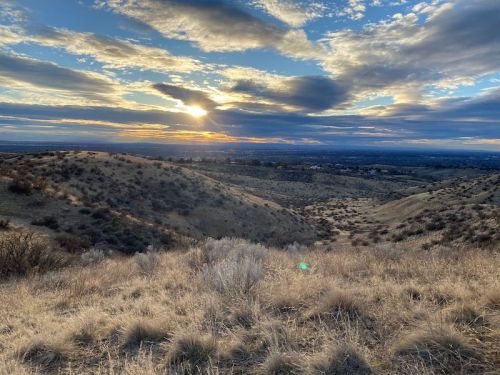 A scenic view of rolling hills under a dramatic sky at sunset, with soft clouds and golden light illuminating the landscape.