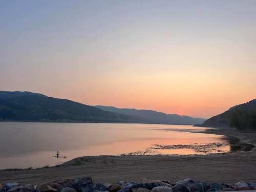 A serene lake at sunset, with mountains in the background and a calm shoreline reflecting the soft colors of the sky.