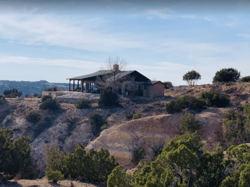 A rustic house perched on a rocky hillside, surrounded by sparse vegetation and a clear blue sky.