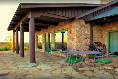 A stone house with large windows, a patio, and outdoor seating, set against a scenic landscape at sunset.