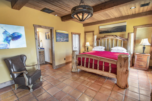 Cozy bedroom with a wooden bed, red bedding, rustic decor, and a leather chair in a warm, inviting atmosphere.