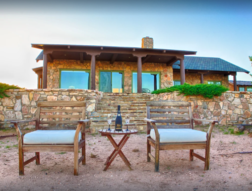 A cozy outdoor seating area with two chairs and a small table, featuring a bottle of champagne and glasses, in front of a stone house.