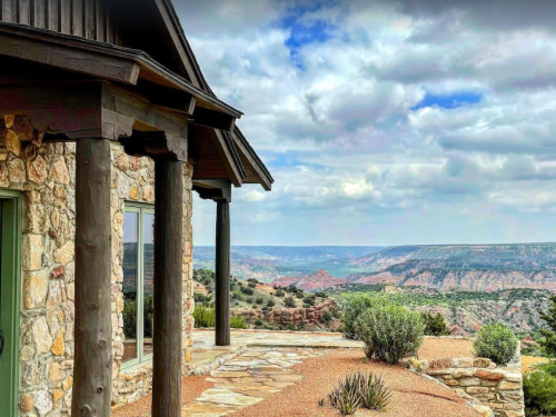 A stone house with wooden beams overlooks a vast canyon landscape under a cloudy sky.