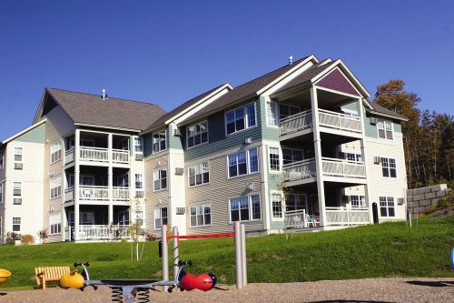 A multi-story apartment building with balconies, surrounded by green grass and a playground in the foreground.