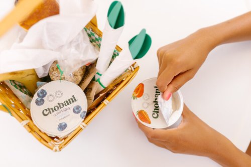 A person opening a Chobani yogurt container next to a picnic basket filled with snacks and utensils.