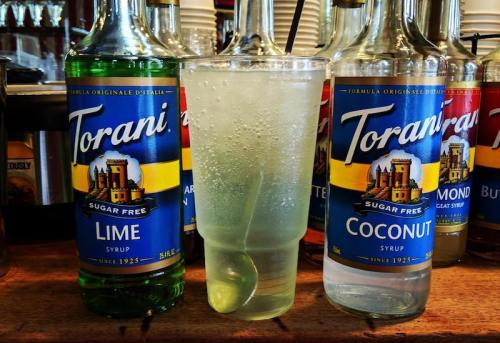 A clear cup filled with a fizzy drink, next to bottles of Torani sugar-free lime and coconut syrup on a bar counter.