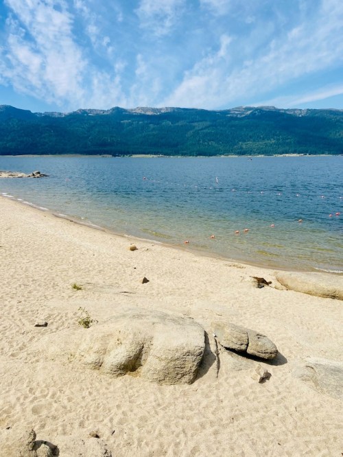 A serene beach scene with sandy shore, calm water, and mountains in the background under a blue sky with wispy clouds.