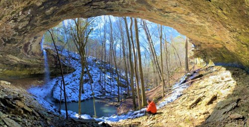 A person in an orange jacket sits inside a cave, overlooking a snowy landscape with a waterfall and trees.