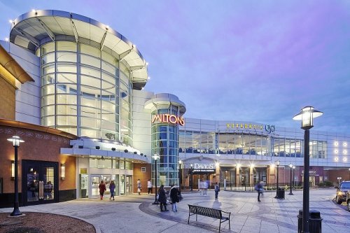 A modern shopping mall entrance with bright signage and people walking outside during twilight.