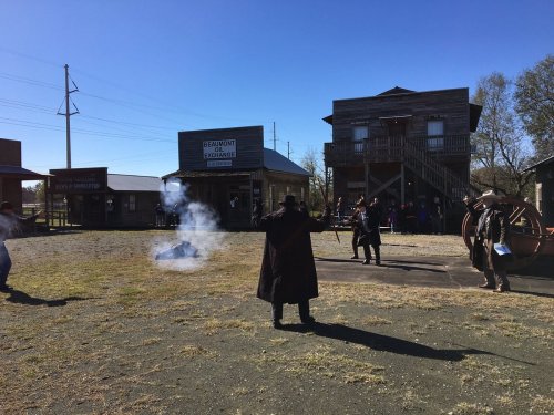A western-style town with buildings, people in period costumes, and a smoke effect, suggesting a staged gunfight.