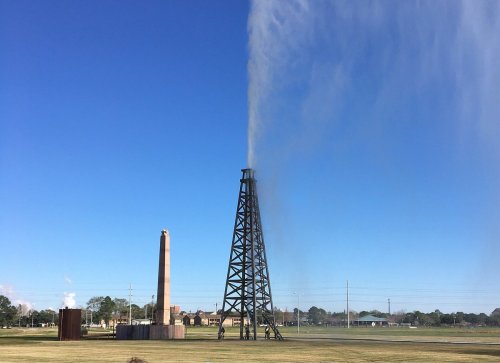 A tall oil rig with water spraying into the air, set against a clear blue sky and grassy landscape.
