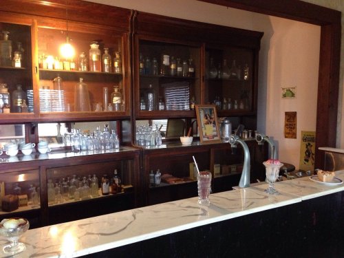 A vintage soda fountain with glassware, bottles, and a dessert display in a wooden cabinet setting.