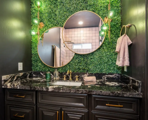 A stylish bathroom with a dark marble countertop, two round mirrors, green wall decor, and modern lighting fixtures.
