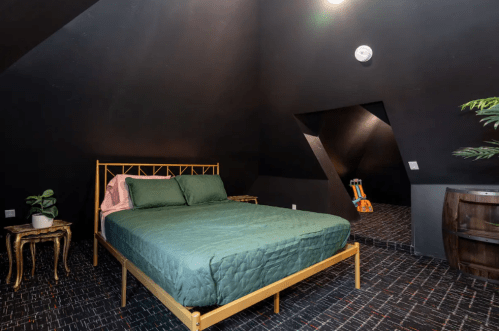 Cozy bedroom with a green bedspread, gold bed frame, and dark walls, featuring a plant and a barrel table.