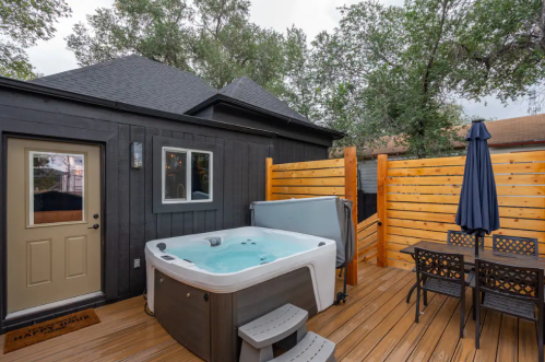 A cozy outdoor space featuring a hot tub, wooden deck, dining table, and a privacy fence surrounded by trees.