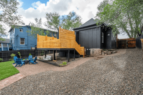 A backyard with a wooden deck, gravel path, and blue chairs, surrounded by trees and a black house.