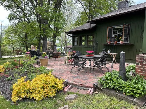 A cozy patio with black metal furniture, colorful flowers, and lush greenery, surrounded by trees and a charming house.
