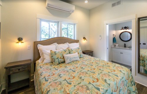 Cozy bedroom with a patterned quilt, bedside tables, and a mirror, featuring natural light from windows.