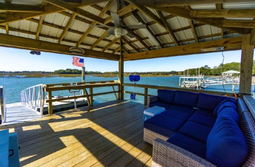 A wooden deck with a cozy seating area overlooking a calm waterway and a flag waving in the breeze.