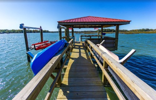 A wooden dock extends over water, with kayaks resting on the side and a red-roofed gazebo in the background.