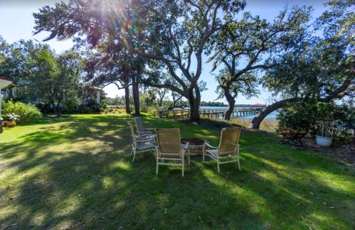 A serene outdoor space with chairs arranged around a fire pit, surrounded by trees and a view of a pier.