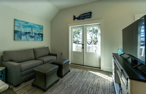 Cozy living room with a gray sofa, two ottomans, a TV, and a view of outdoor scenery through double doors.