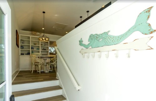 A staircase leads to a bright room with a mermaid wall decoration and a dining area in the background.