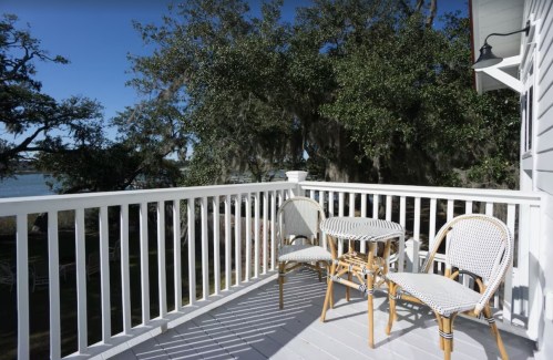 A sunny balcony with two wicker chairs and a small table, surrounded by trees and a view of the water.