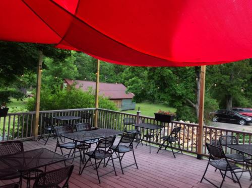 Outdoor patio with black tables and chairs, shaded by a large red canopy, surrounded by greenery and a rustic building.