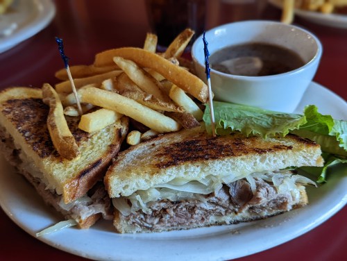 A hearty sandwich with pulled meat and sauerkraut, served with fries and a small bowl of soup on a plate.
