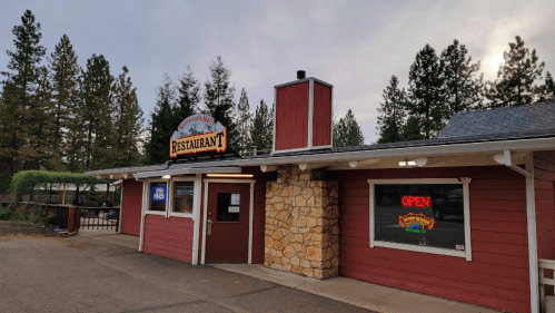 A rustic restaurant with a stone facade and a sign reading "Open," surrounded by tall trees and a cloudy sky.