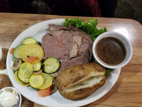 A plate with roast beef, a baked potato, sautéed vegetables, and a small bowl of gravy, garnished with lettuce.