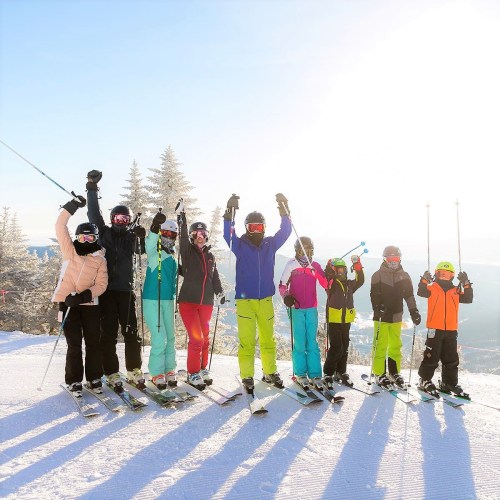 A group of eight skiers in colorful gear poses joyfully on a snowy slope, with trees and sunlight in the background.